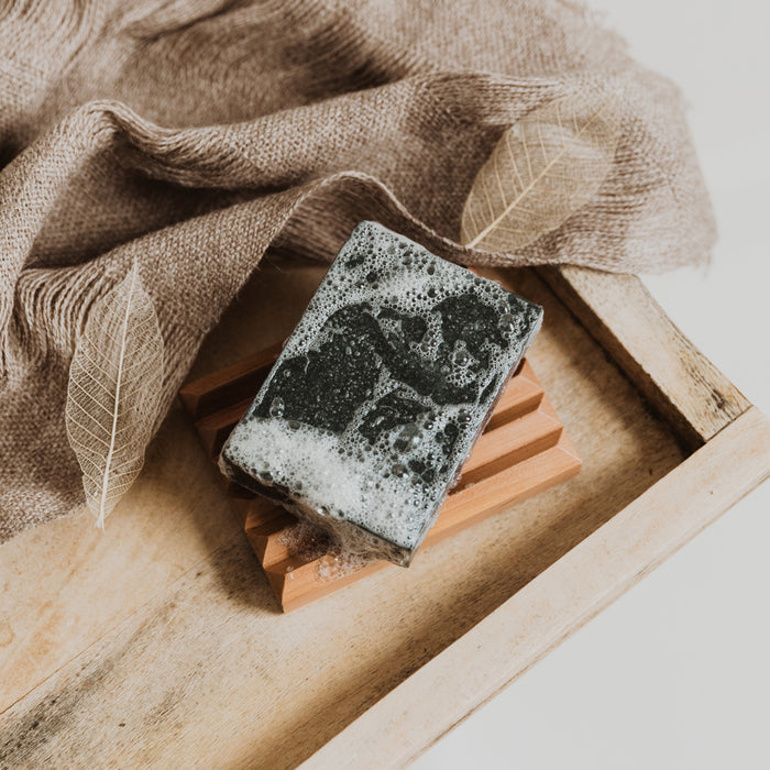 The Activated Charcoal Facial & Body Bar by Little Seed Farm rests on a wooden soap dish atop a rustic tray, accented with a textured brown cloth and two decorative skeleton leaves.