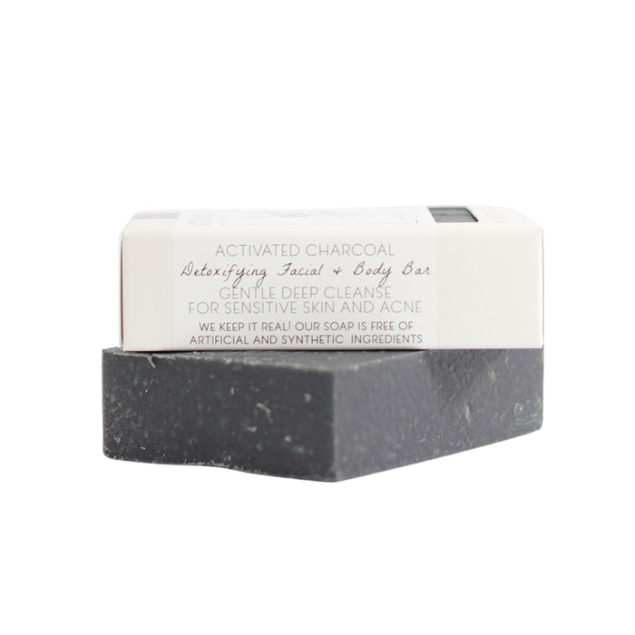 A black rectangular Activated Charcoal Facial & Body Bar by Little Seed Farm, partially wrapped in white packaging labeled for sensitive and acne-prone skin, rests on a plain white background.