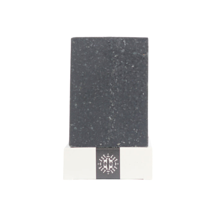 The Little Seed Farm Activated Charcoal Facial & Body Bar, a rectangular dark stone bar for sensitive or acne-prone skin, stands upright on a white base with a black band and snowflake pattern, set against a plain white background.