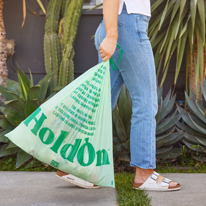 A person carrying HoldOn Bags Compostable Tall Kitchen Bags, 13 gal – Unscented, outside is dressed in blue jeans, a white top, and white sandals, with plants and a cactus in the background.