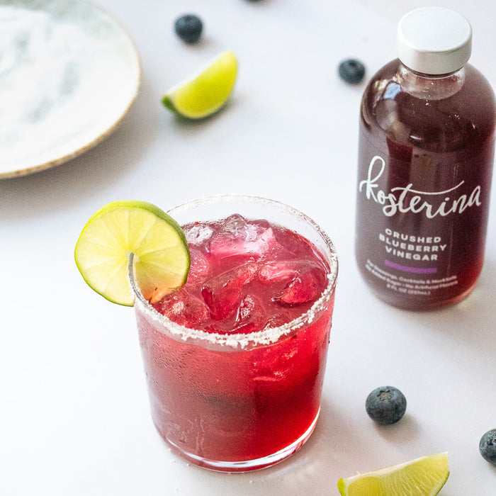 A glass of red iced drink with a salted rim and lime slice sits on a white surface next to Kosterina Crushed Blueberry Vinegar, a fruit vinegar with no added sugar, plus lime wedges, blueberries, and a plate.