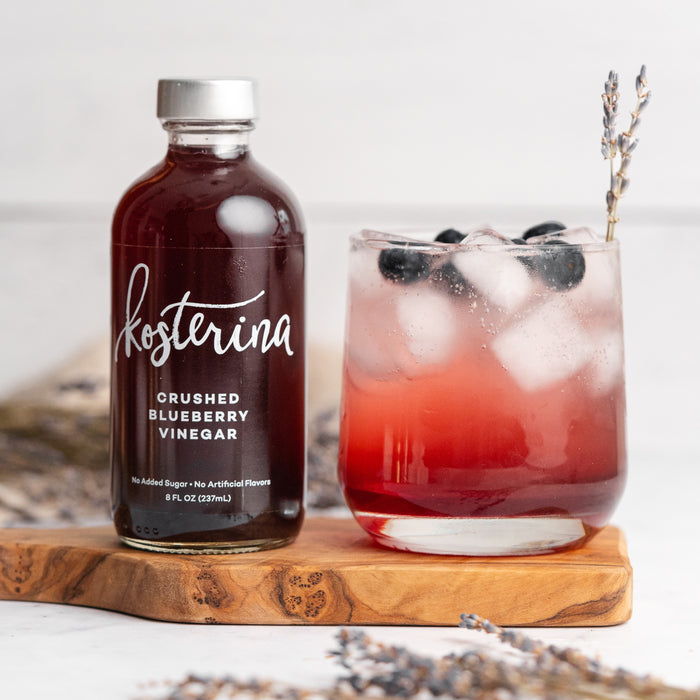 A bottle of Kosterina Crushed Blueberry Vinegar sits beside a glass of iced red drink garnished with blueberries and lavender on a wooden board, set against a blurred floral background.