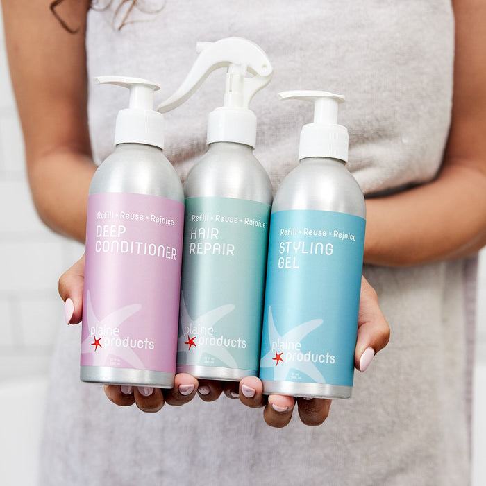 A person holds Plaine Products Deep Conditioner and two styling products in sleek silver bottles with colorful labels—ideal for hydrating hair, restoring shine, and nourishing as a hair moisturizer.