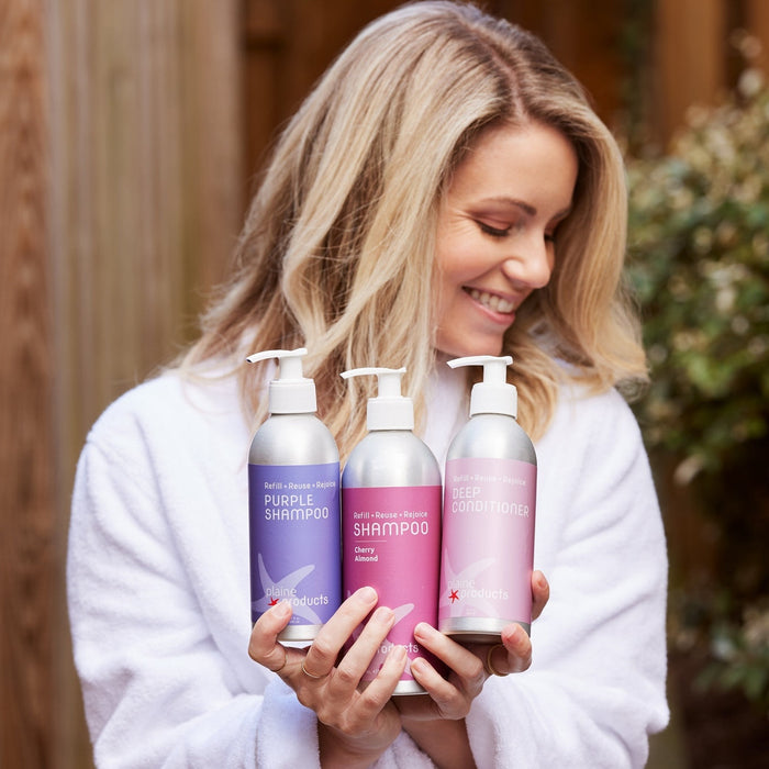 A smiling woman in a white robe holds Plaine Products’ Deep Conditioner, standing outside with greenery behind her, ready to restore shine to her hair.