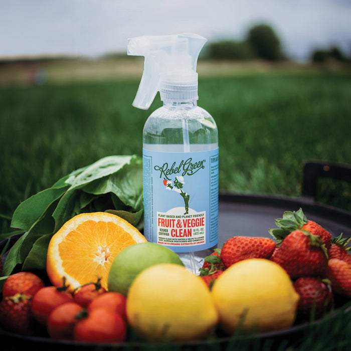A bottle of Rebel Green Fruit & Veggie Clean Produce Wash Spray rests on a tray with fresh lemons, strawberries, orange, and leafy greens outdoors, ready to naturally remove pesticides from your produce.