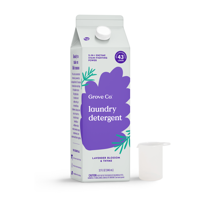 A carton of Grove Co. Laundry Detergent – 5-in-1 Enzyme Stain Fighting Power with a new purple label and green accents is placed beside a clear measuring cup. It delivers 43 loads and features a lavender blossom scent.