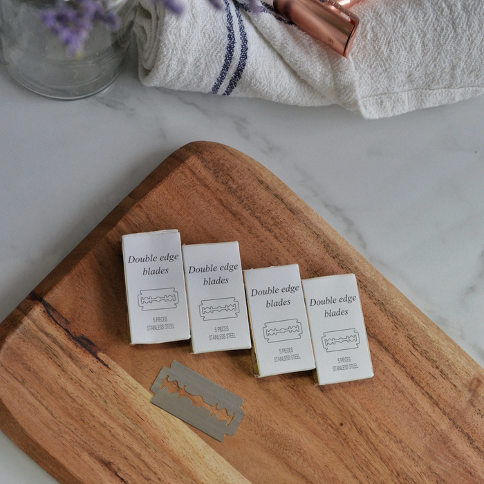 Four Me Mother Earth Safety Razor Refill Pack boxes and two loose blades are arranged on a wooden board, with a towel, lipstick, and a jar of purple flowers in the background on a marble surface.