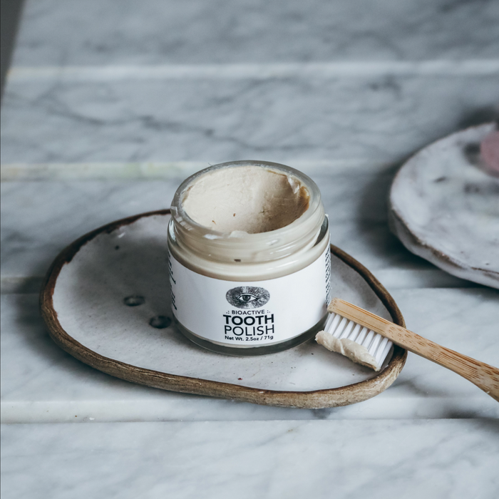 Anima Mundi's TOOTH POLISH, Fluoride Free, is displayed open in a glass jar on a ceramic dish beside a bamboo toothbrush with white bristles—all atop marble—a chic, plant-based oral care solution.